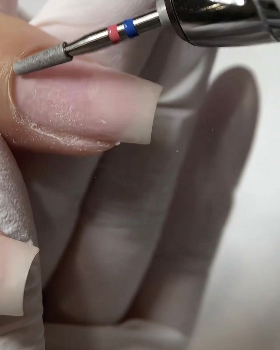 Un primer plano de una fresa de cilindro plano de torno exfoliando y empujando suavemente la zona de la cutícula proximal en una uña natural.