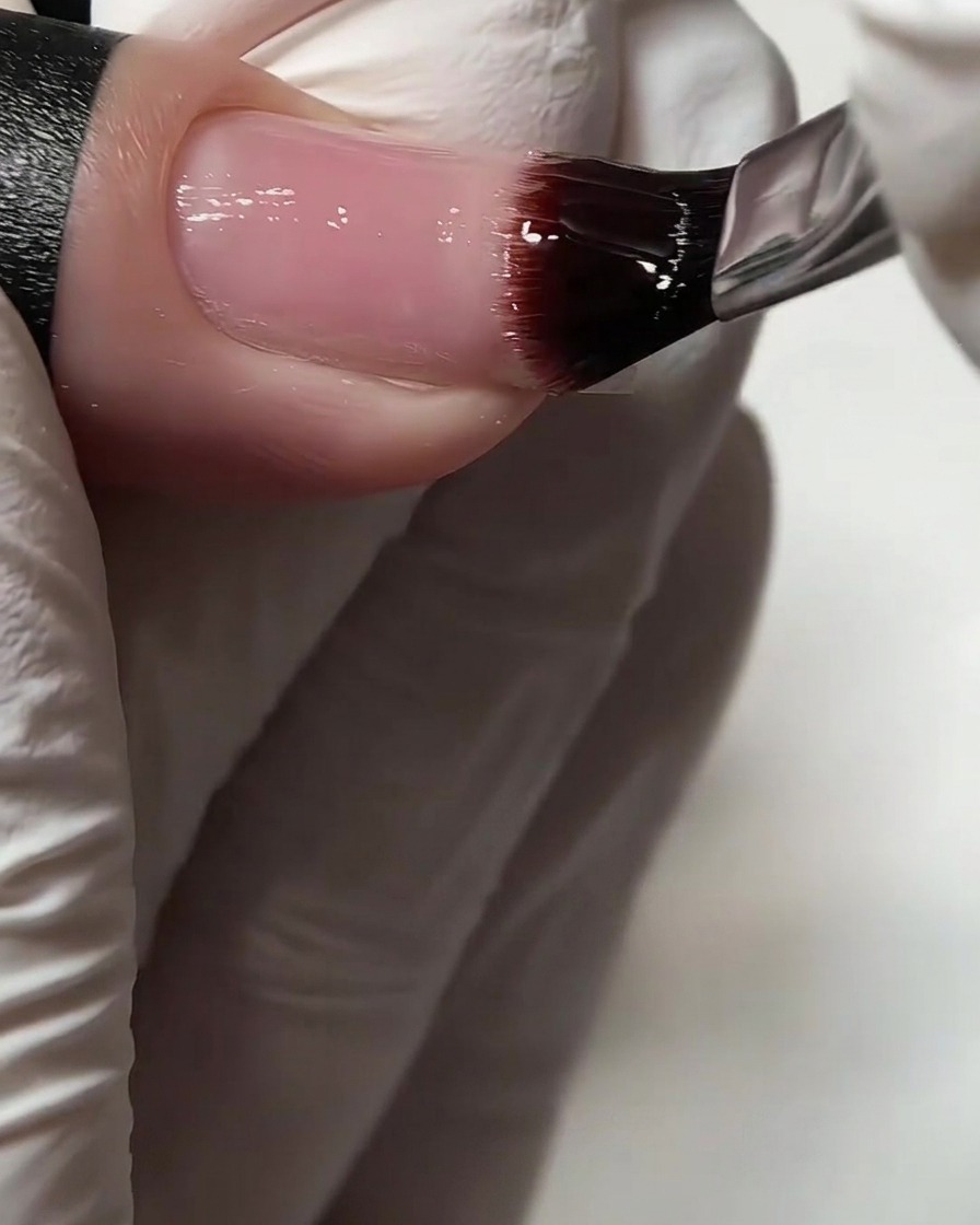 Un primer plano de un pincel de gel plano aplicando una capa base transparente sobre una uña natural.