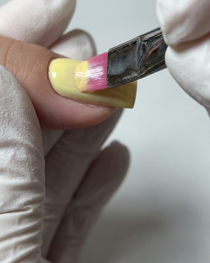 Un primer plano de un pincel aplicando una segunda capa de esmalte en gel amarillo pastel para asegurar una cobertura de color completa.