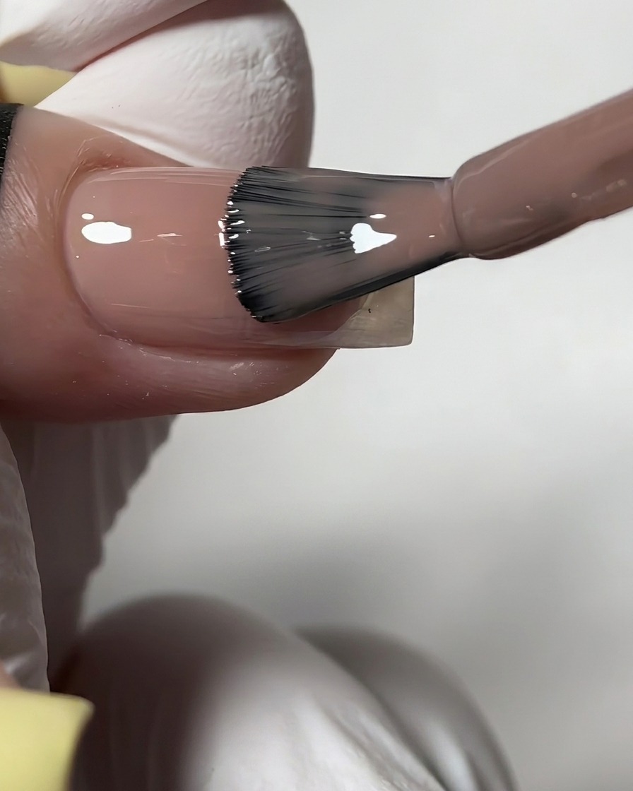 Un pincel de botella aplicando un esmalte en gel nude translúcido y brillante sobre una uña preparada.