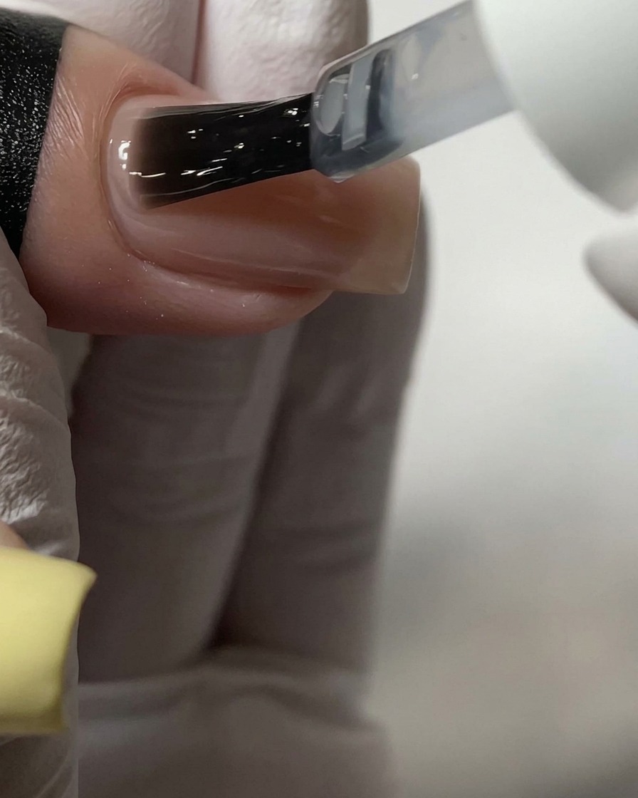 Un pincel aplicando una capa superior transparente sobre una uña acrílica de color nude.
