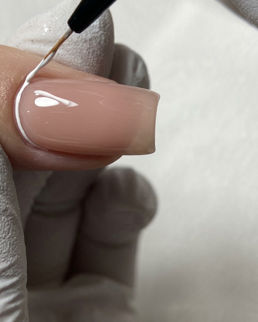 Un pincel de nail art fino dibujando líneas geométricas blancas cerca de la cutícula en una uña nude.