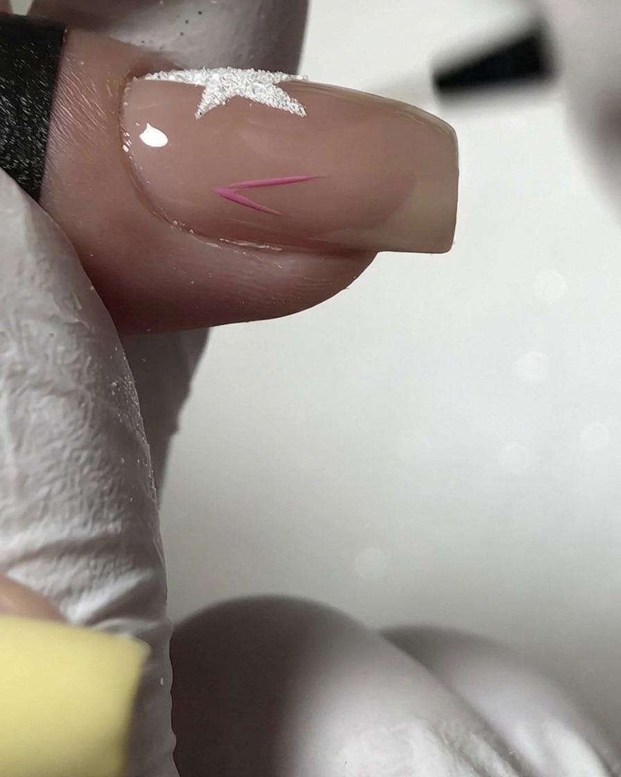 Un pincel fino pintando el contorno de una pequeña estrella rosa en la sección inferior de una uña nude.