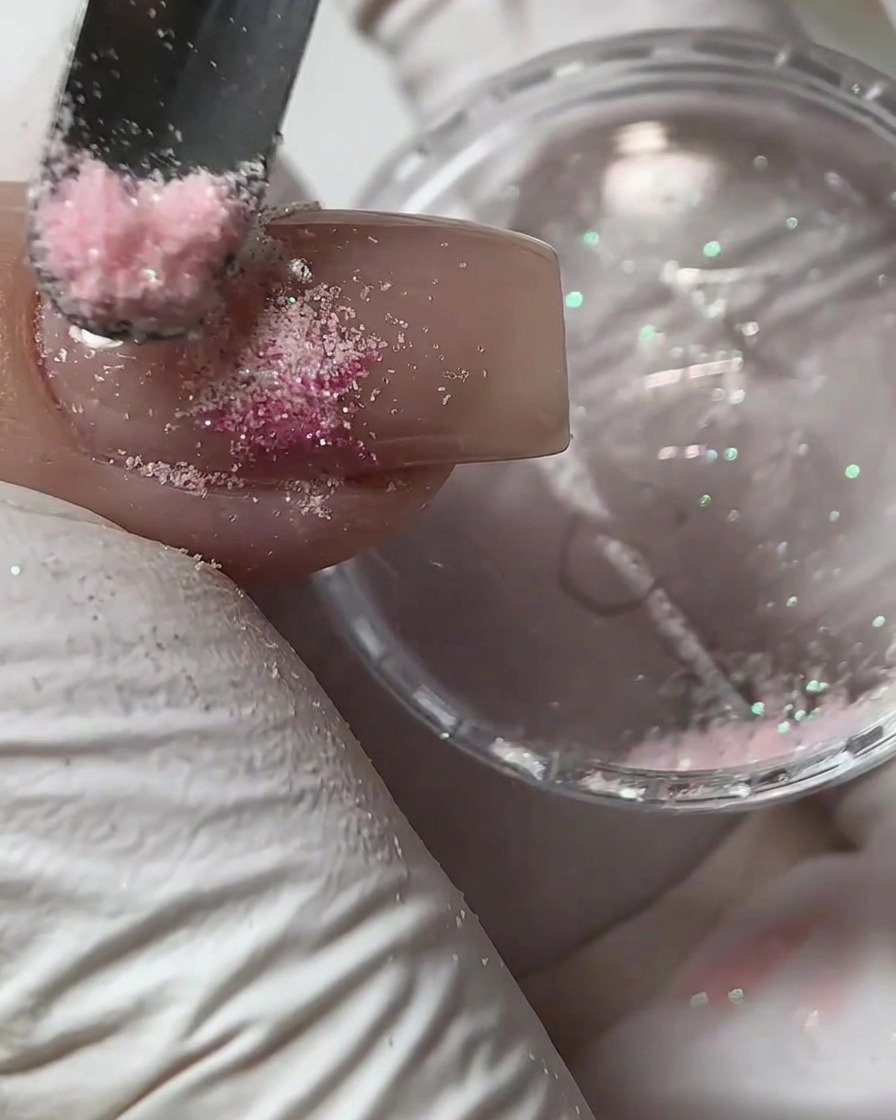 Una pequeña herramienta esparciendo polvo de azúcar rosa sobre un diseño de gel húmedo en una uña.