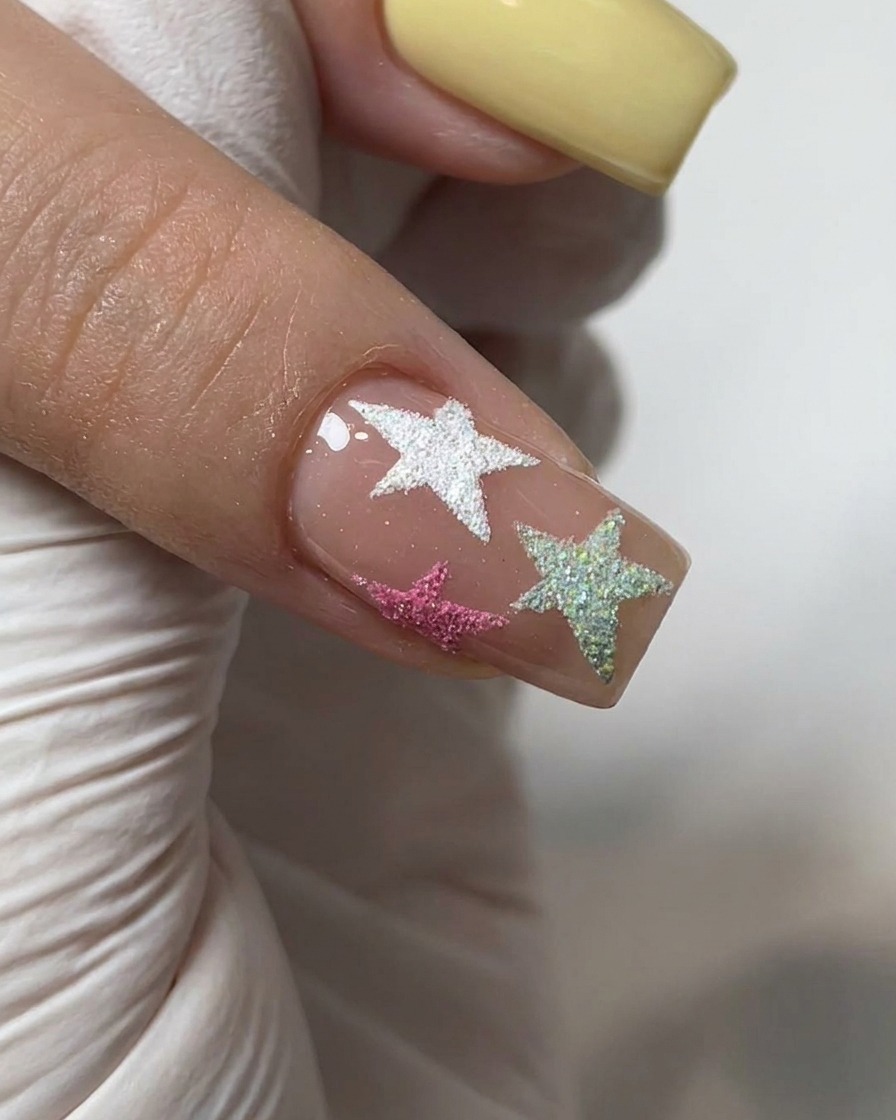 Un pincel aplicando una capa superior transparente y brillante sobre una uña decorada con polvo de cromo y estrellas texturizadas.