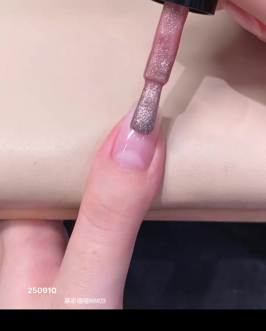 Un pincel de esmalte aplicando una gota de esmalte en gel magnético rosa translúcido cerca del área de la cutícula de una uña natural.