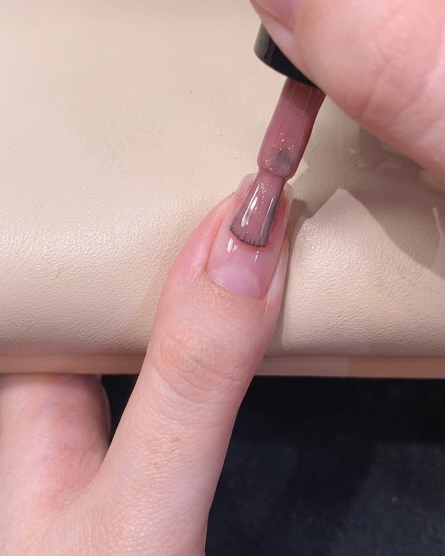 Un pincel de esmalte en gel deslizándose por el centro de la uña, distribuyendo un esmalte brillante de color rosa translúcido.