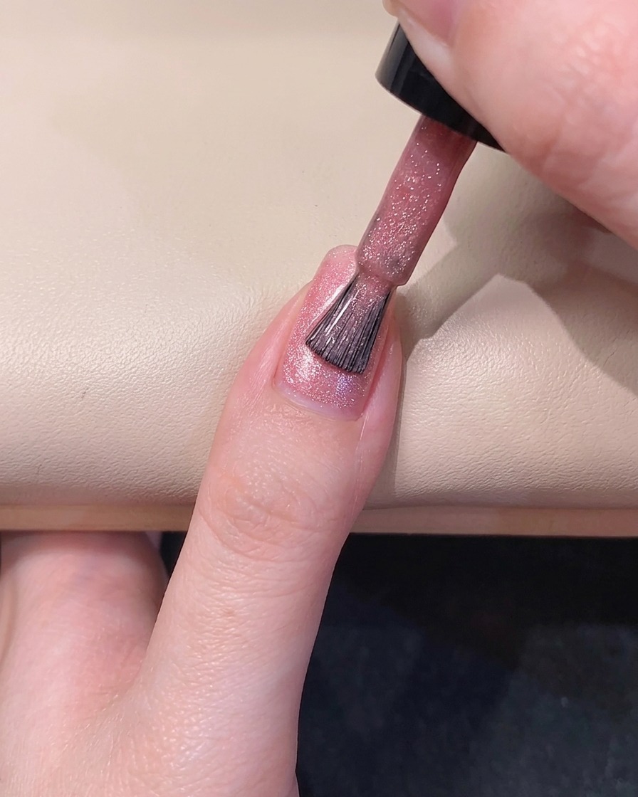 Un pincel de uñas esparciendo esmalte en gel rosa translúcido a lo largo del lado izquierdo de la uña.