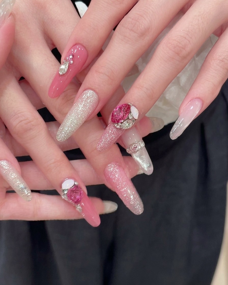 Dos manos perfectamente manicuradas descansando sobre un fondo de tela oscura, mostrando uñas largas y rosadas en forma de almendra con degradados de glitter plateado y grandes acentos de pedrería de corazón rosa.