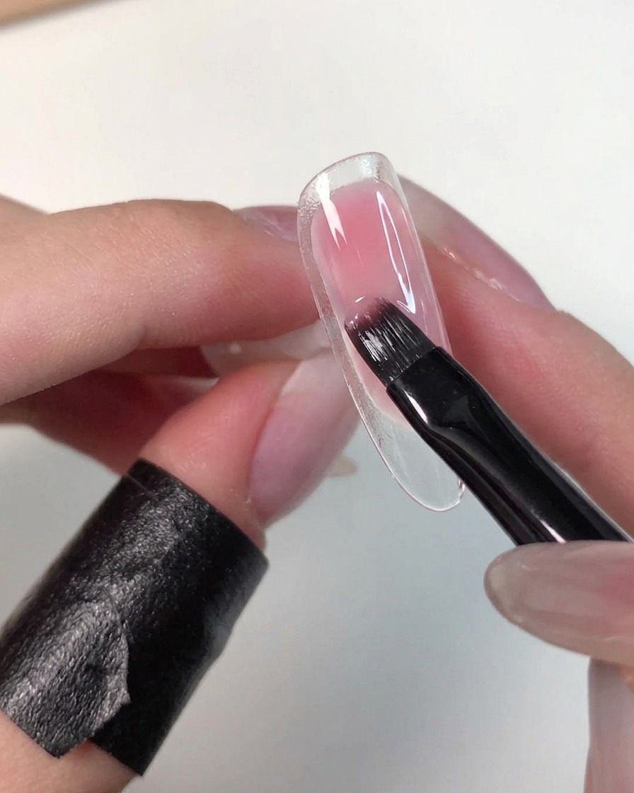 Una mano sosteniendo una punta de uña transparente mientras un pincel plano difumina suavemente el gel rosa húmedo para suavizar sus bordes.