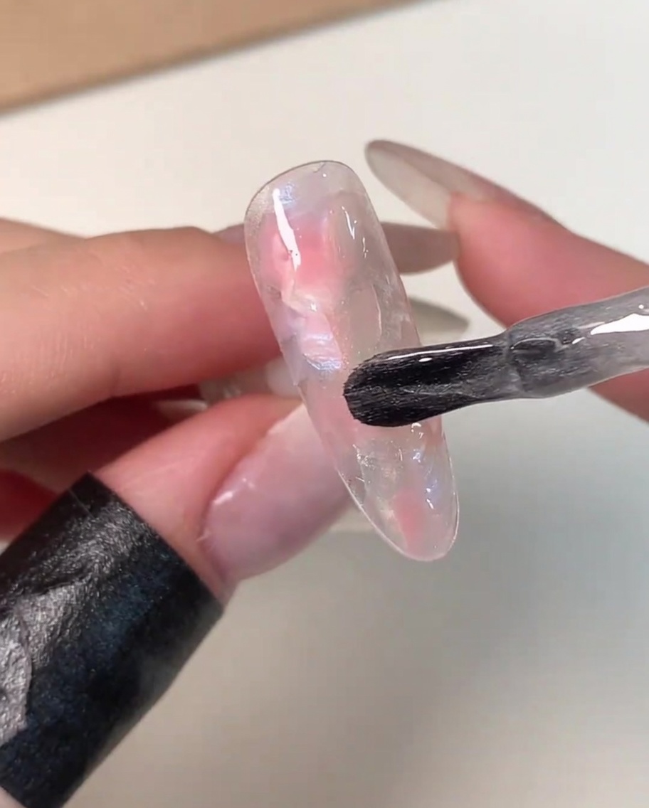 Un pincel de botella aplicando una capa de gel magnético transparente y brillante sobre una punta de uña marmoleada en rosa y blanco.