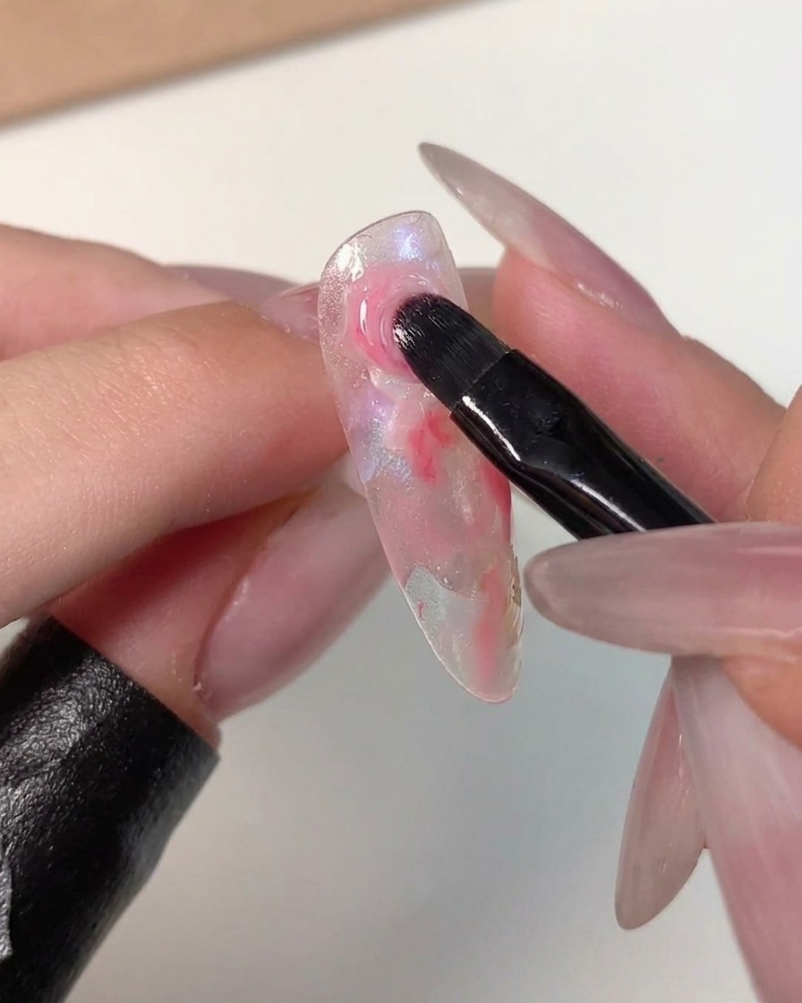 Un pincel de arte de uñas difuminando los bordes de un punto rosa oscuro hacia los colores más claros circundantes en una punta de uña transparente.
