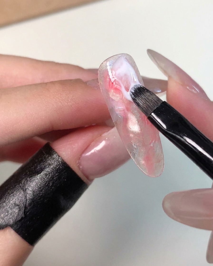 Un pincel de gel pequeño aplicando un trazo de gel blanco lechoso cerca del área de la cutícula de una punta de uña marmoleada para refinar el patrón.