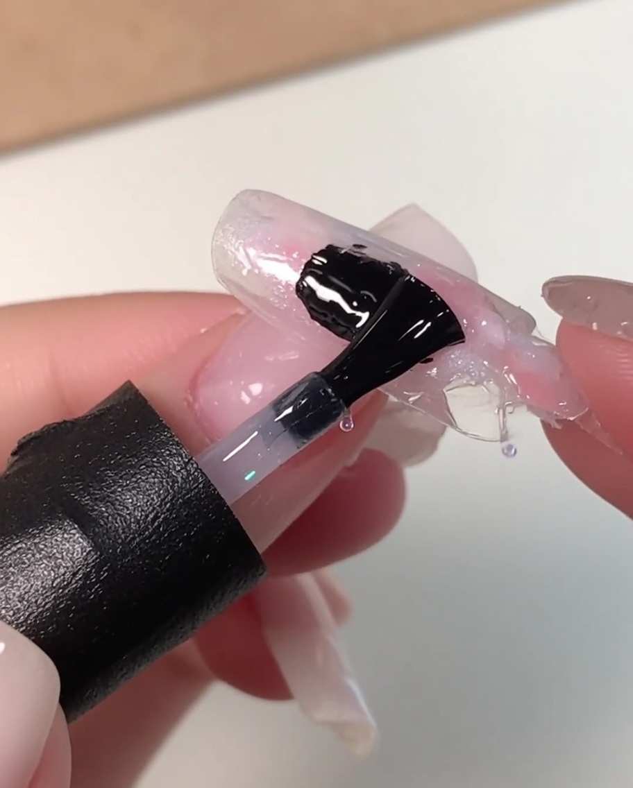 Un pincel de uñas negro aplicando una capa gruesa de gel nivelador transparente sobre un diseño de uñas marmoleado en rosa, blanco y azul.