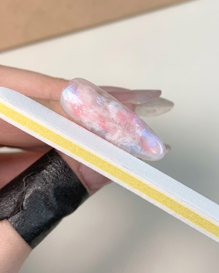 Una mano sosteniendo una tira de lima de uñas blanca y amarilla, puliendo la superficie de una uña de gel marmoleada, curada y brillante.