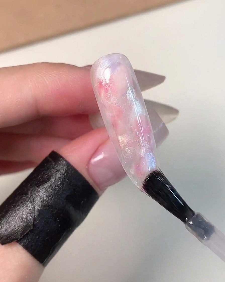 Un pincel de uñas aplicando una capa superior transparente y brillante sobre una uña con efecto de bola de vidrio marmoleada en rosa, azul y blanco terminada.
