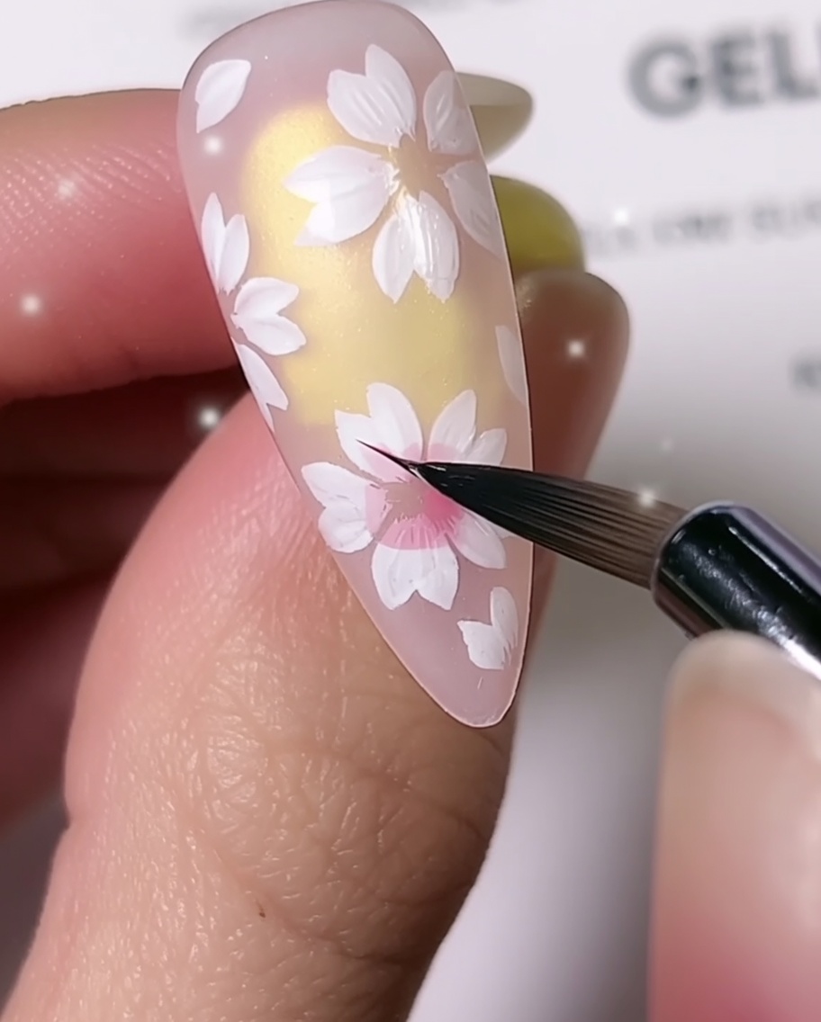 Un primer plano que muestra un pincel fino para detalles agregando sombreado rosa al centro de los pétalos blancos de flor de cerezo en una uña almendradacite: 1.