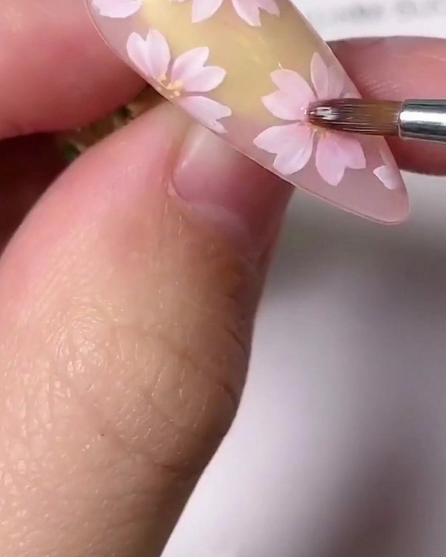 Un primer plano de un pincel de detalles finos aplicando pequeños detalles dorados en el centro de una flor de cerezo rosa pintada en una uña.