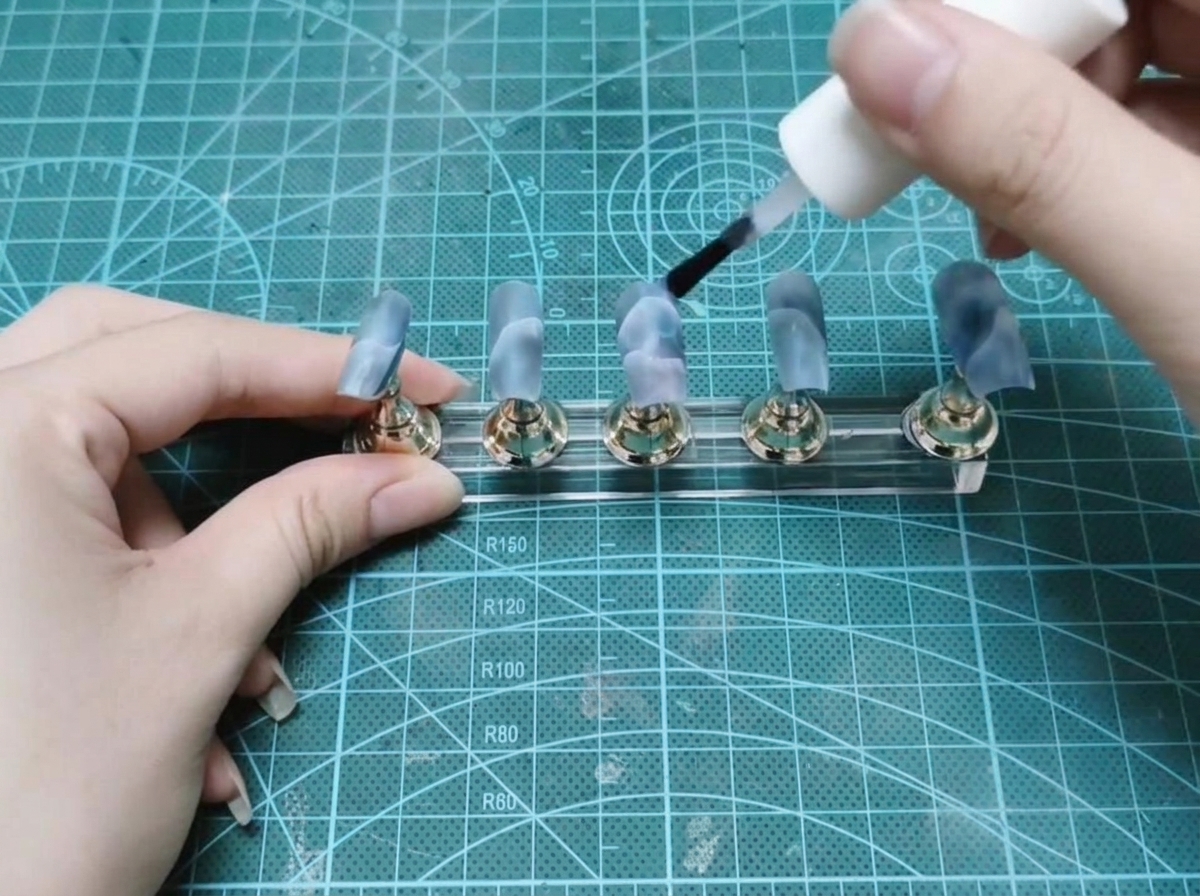Un pincel de botella aplicando gel de construcción transparente a un juego de uñas postizas pintadas en un soporte de exhibición.