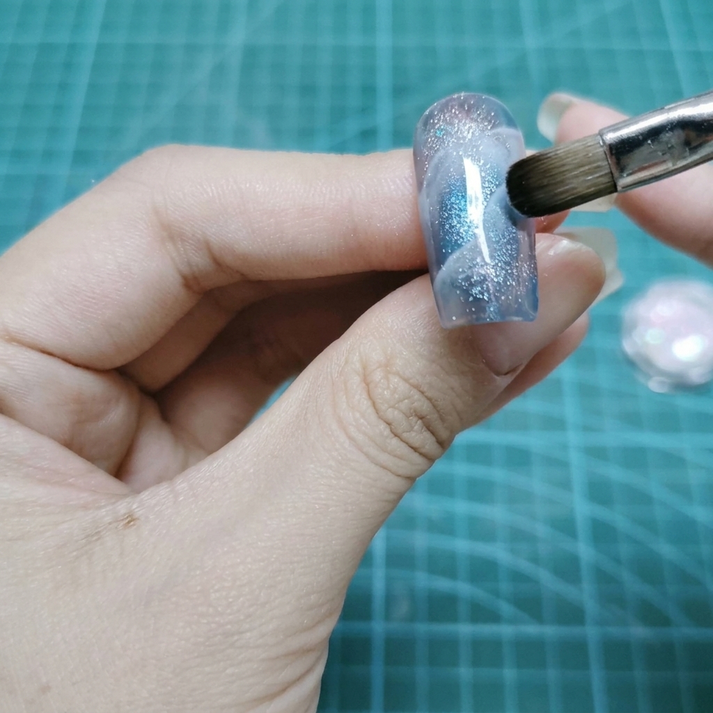 Un pincel de maquillaje plano dando golpecitos con partículas de purpurina holográfica sobre una superficie de uña de gel húmedo.