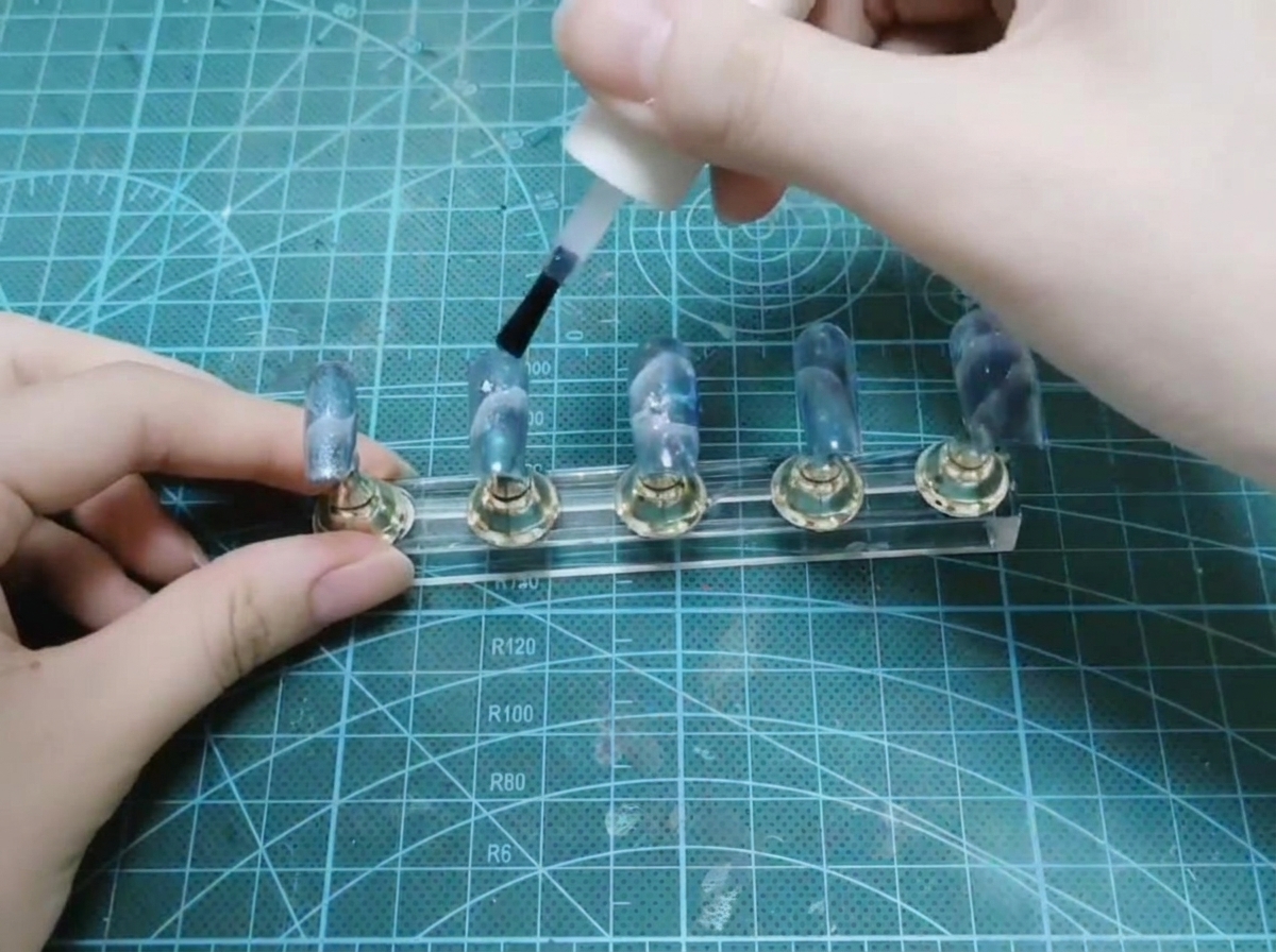 Una mano usando un pincel de una botella blanca para aplicar una capa superior brillante sobre un conjunto de uñas completadas en un soporte de exhibición.