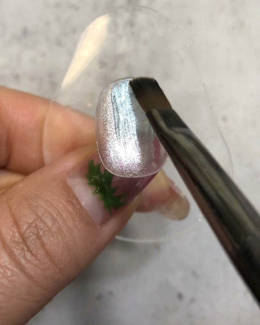 Un pincel de uñas aplicando un esmalte de gel azul traslúcido sobre una base magnética plateada brillante.
