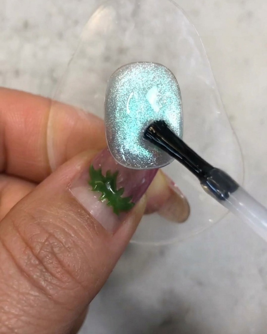 Un pincel aplicando una pequeña gota de gel de refuerzo transparente sobre la superficie texturizada de la uña azul con ojo de gato.