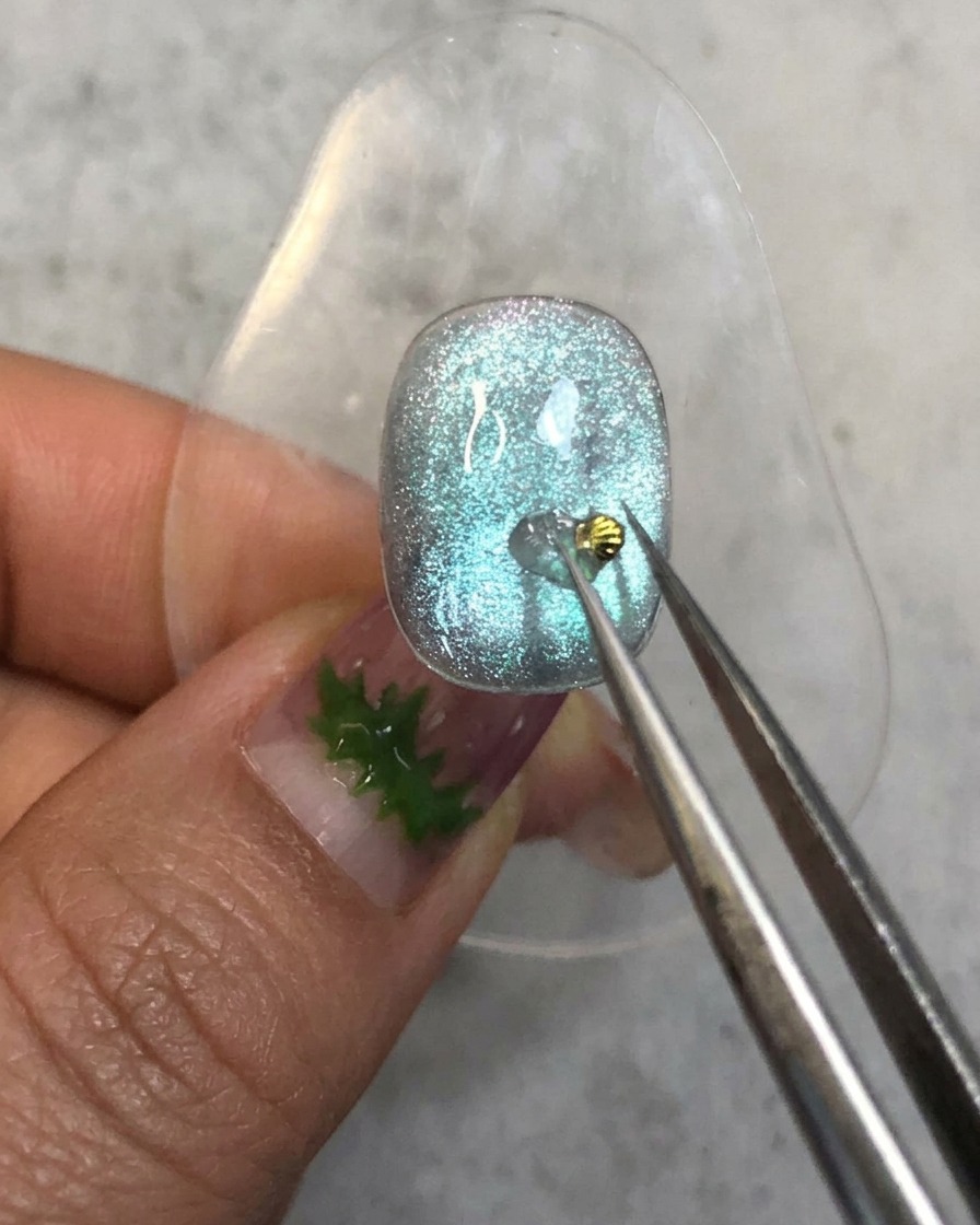 Unas pinzas colocando un diminuto dije de concha dorada sobre el punto de gel de refuerzo en una uña azul.