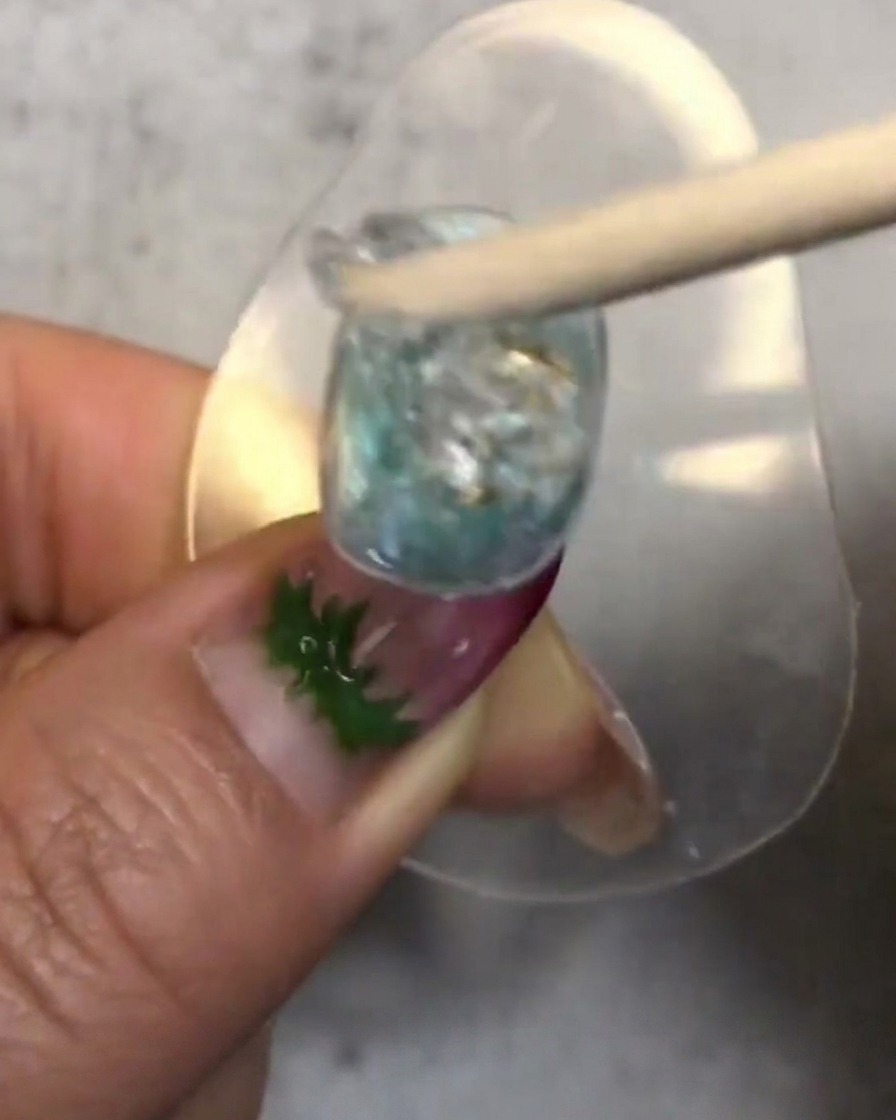 Un palito de madera mezclando y colocando una gota de gel suave candy translúcido sobre la uña decorada.
