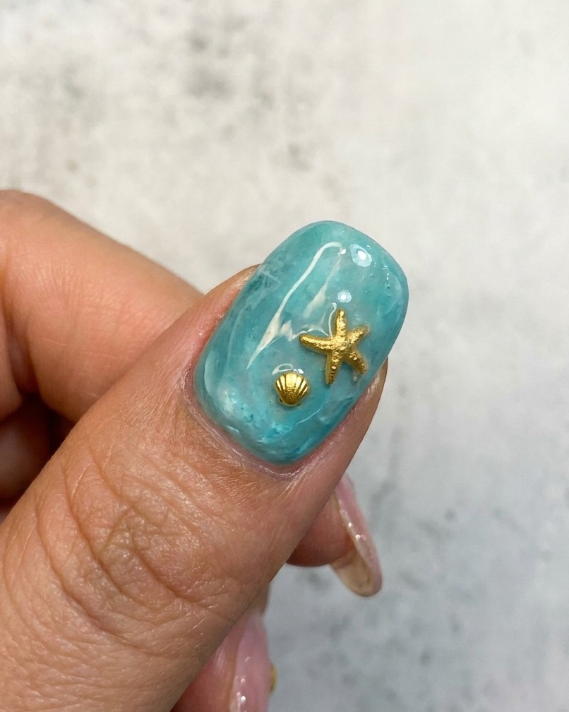 Un primer plano de un diseño de uña oceánica azul con un dije de estrella de mar y concha dorados.