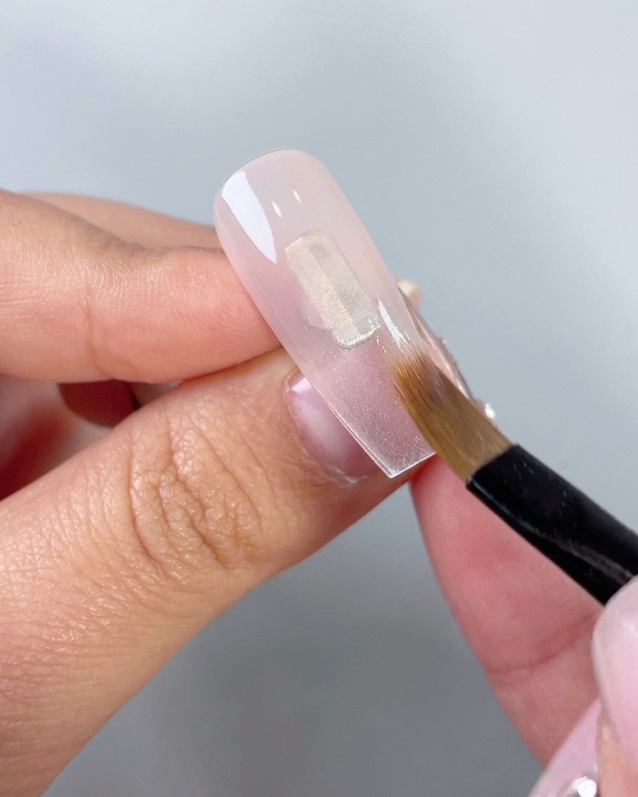 Un primer plano de un pincel de maquillaje plano difuminando gel rosa translúcido hacia abajo en una punta de uña artificial transparente.