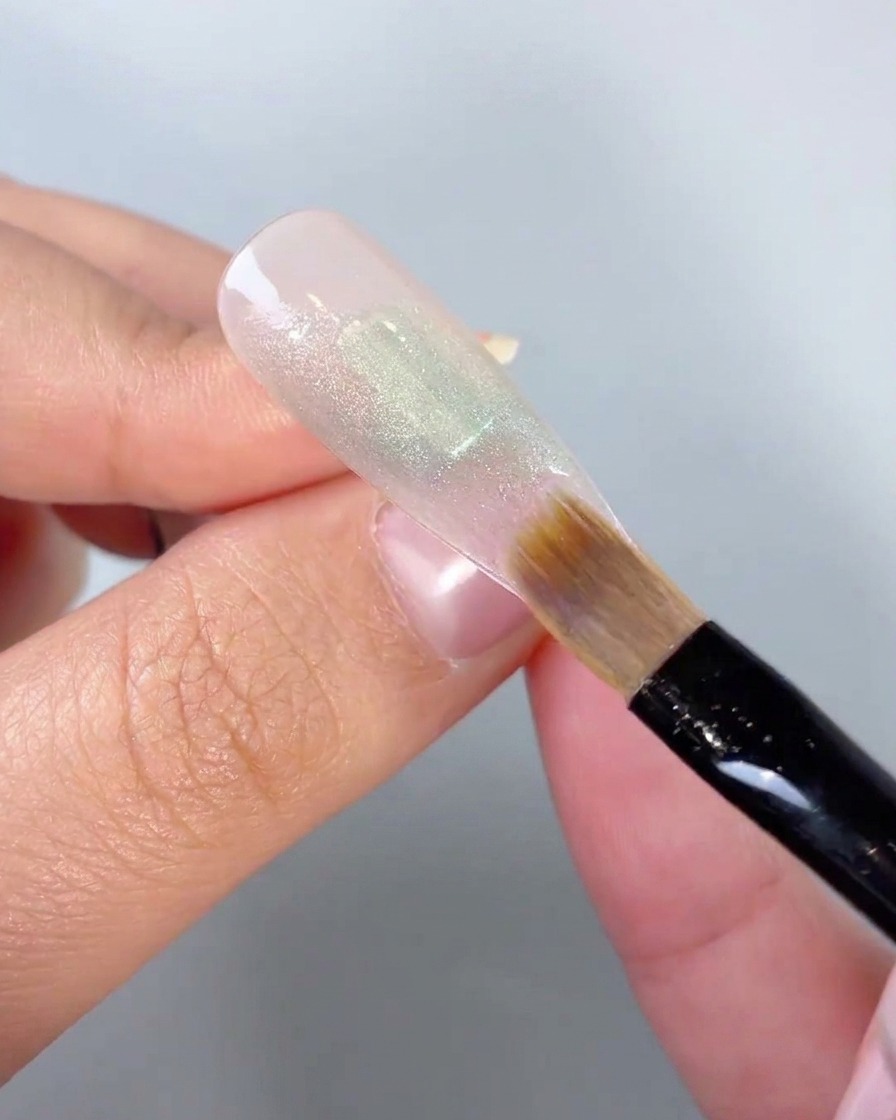 Un primer plano de un pincel suave difuminando los bordes de un bloque de color de gel verde en la punta de una uña.