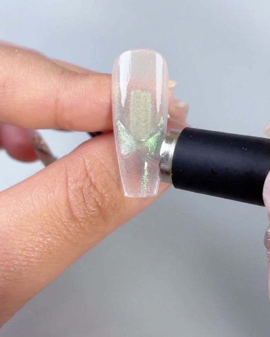 Una varita magnética negra sostenida cerca del costado de una punta de uña transparente que muestra patrones activados de alas de mariposa con ojo de gato verde.
