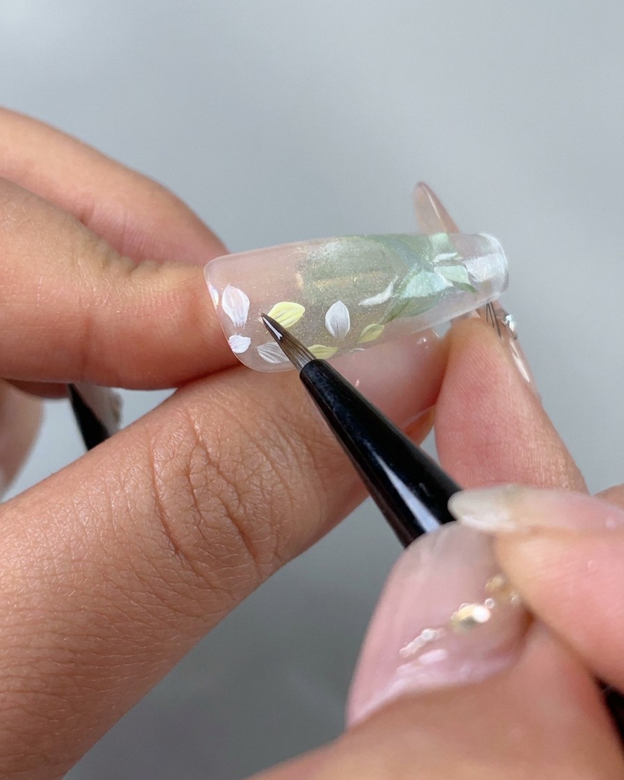Un pincel fino que agrega cuidadosamente tintes de gel rosa claro y amarillo en los pétalos de flores pintados de blanco individuales en la uña.