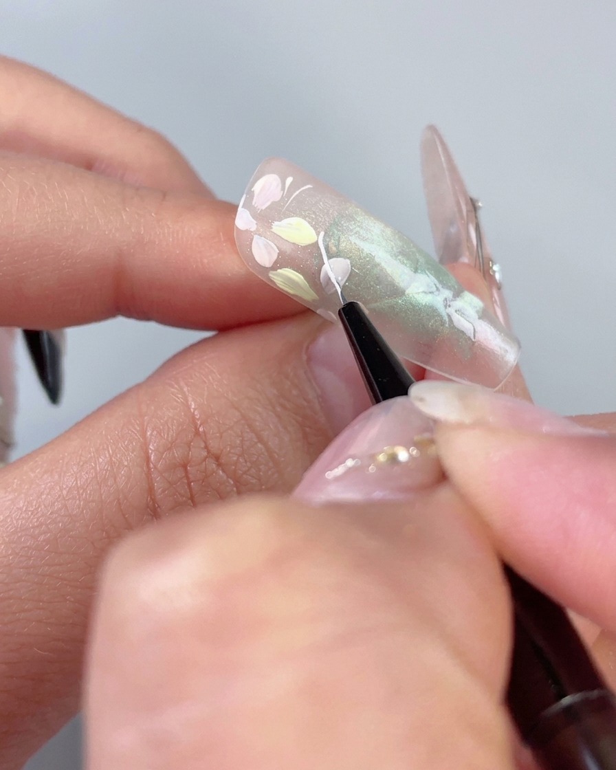 Un pincel delineador fino que dibuja una delicada línea curva blanca al lado de flores pastel en una uña de ojo de gato verde translúcido.