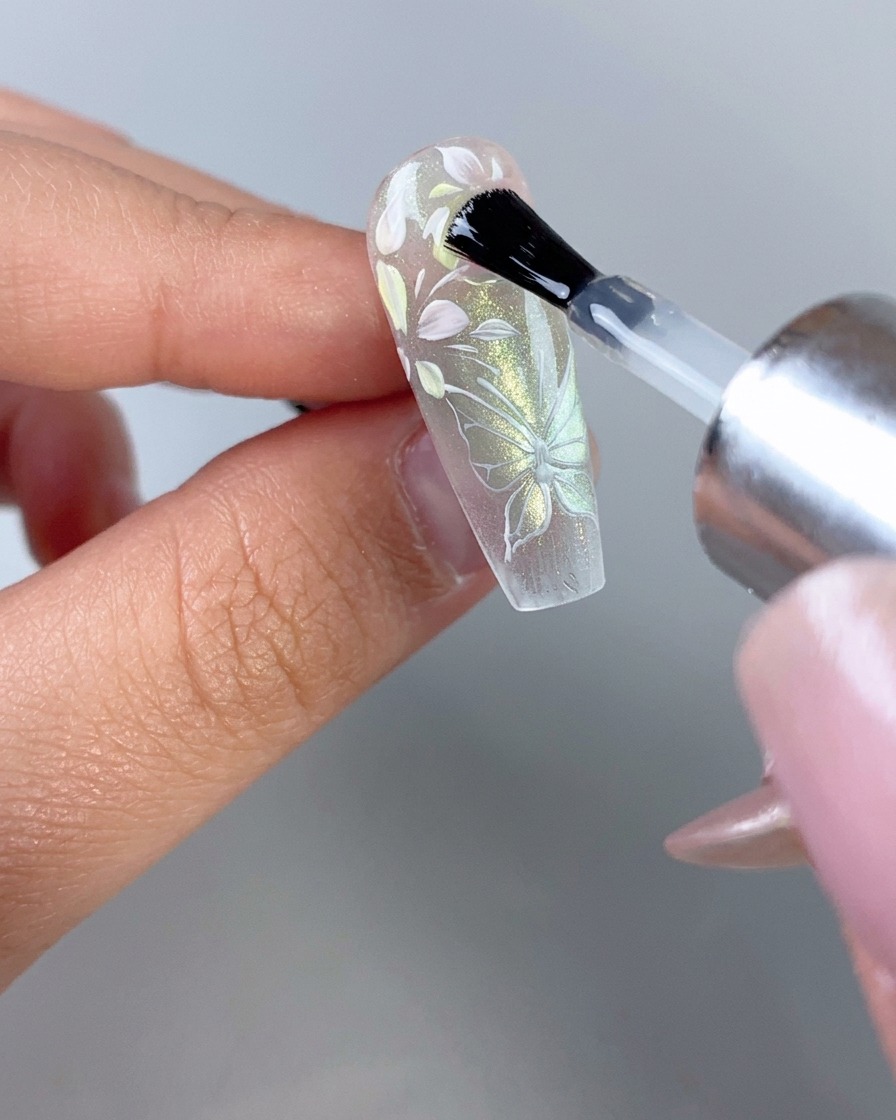 Un pincel de esmalte de uñas aplicando una capa superior transparente brillante sobre una uña totalmente decorada con diseño floral y de mariposa translúcido.