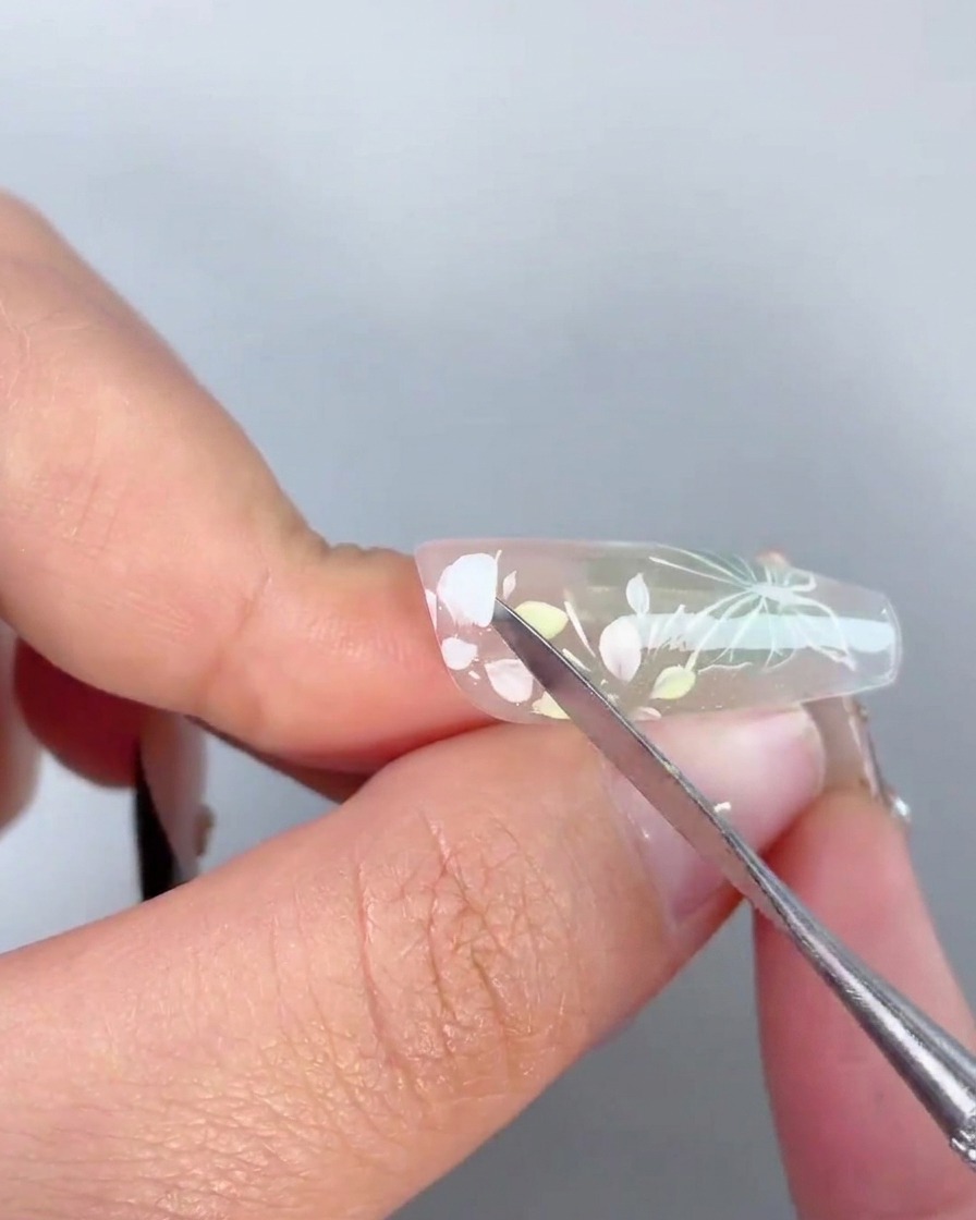 Una herramienta de metal aplicando una gota de gel adhesivo transparente en el centro de un diseño floral pintado en una uña.