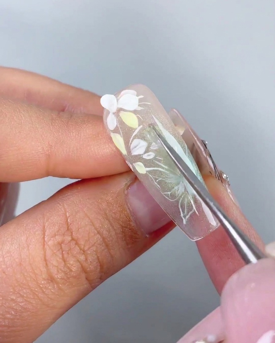 Una herramienta de metal colocando un pétalo blanco 3D preformado sobre el gel adhesivo de una uña.