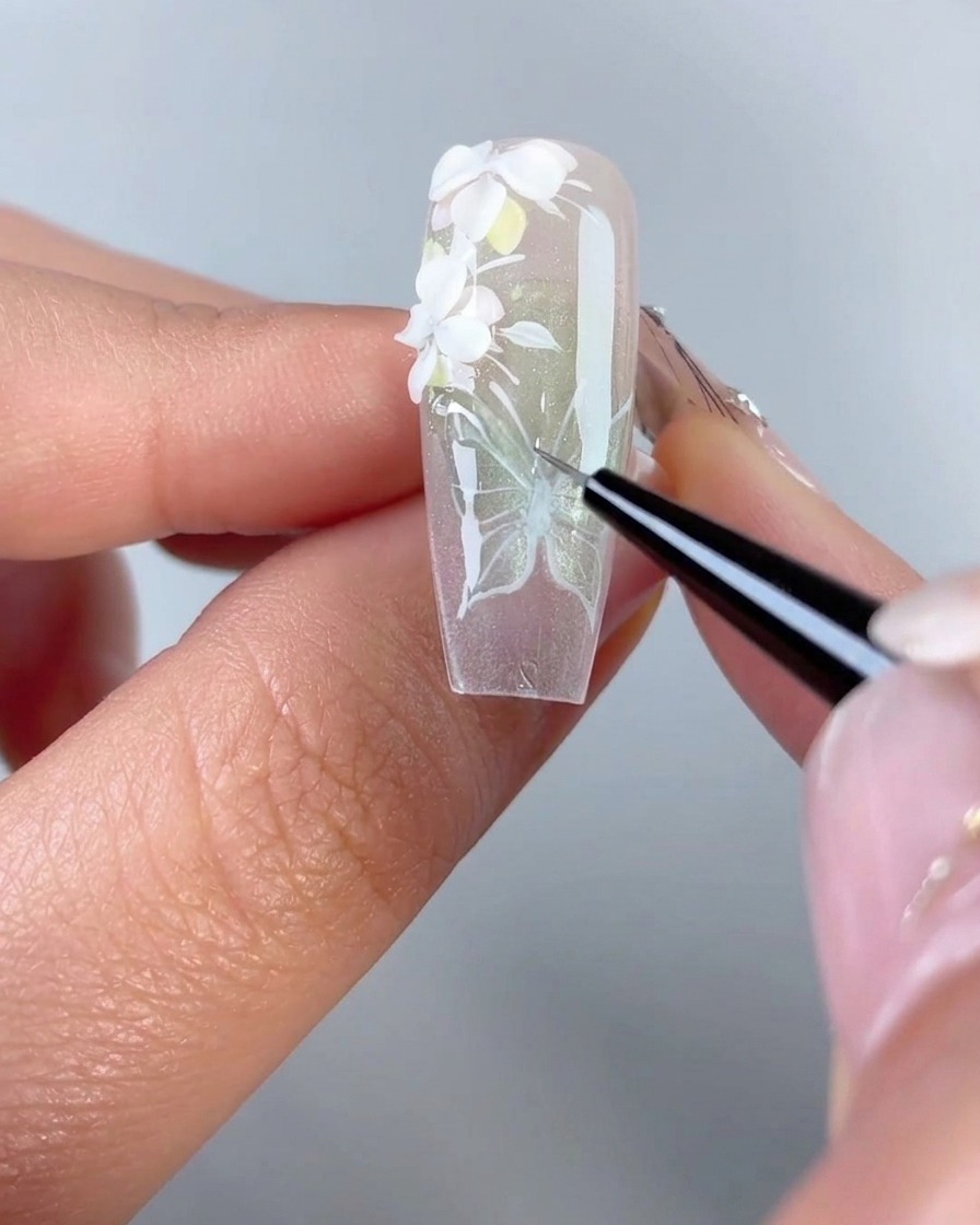 Un primer plano de un pincel delineador fino trazando gel de modelado 3D transparente sobre las líneas blancas pintadas de un diseño de alas de mariposa en la punta de una uña de gel.