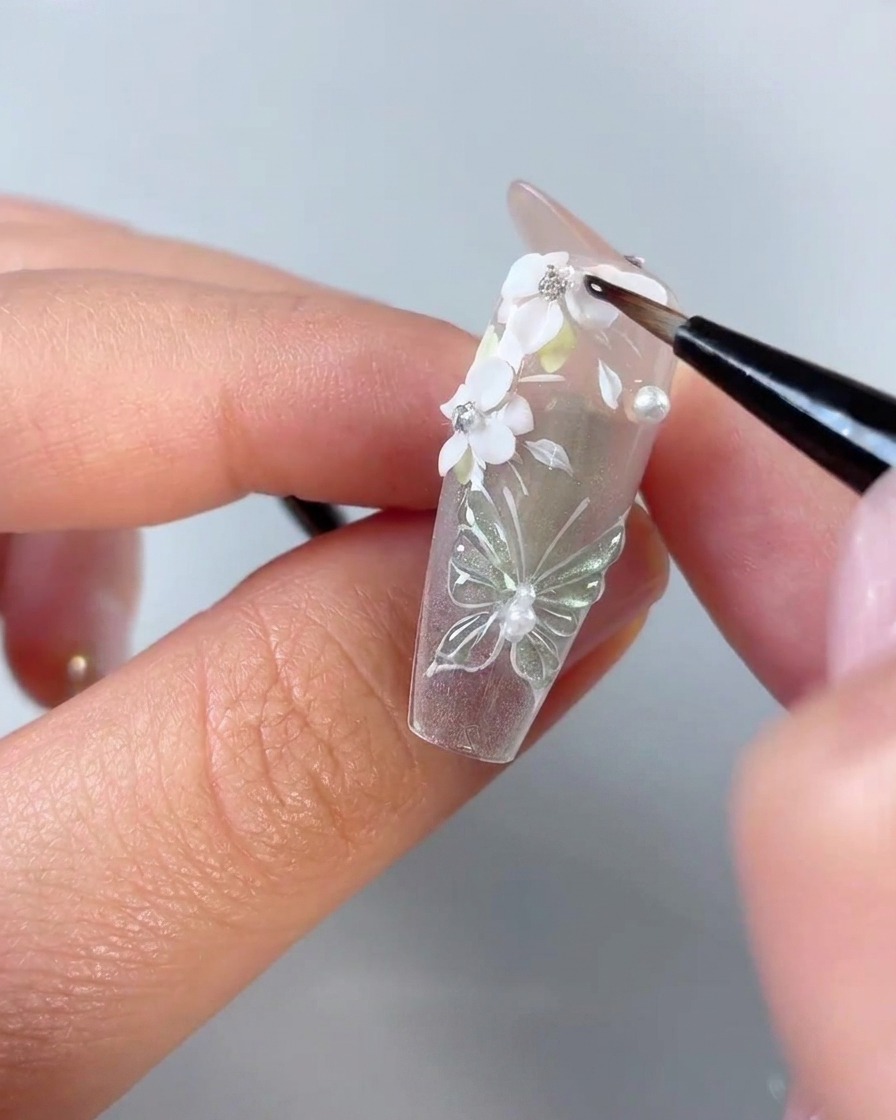 Un pincel de detalle fino para uñas aplicando gel transparente alrededor de los bordes de los pétalos de flores blancas 3D y pedrería en una punta de uña transparente.