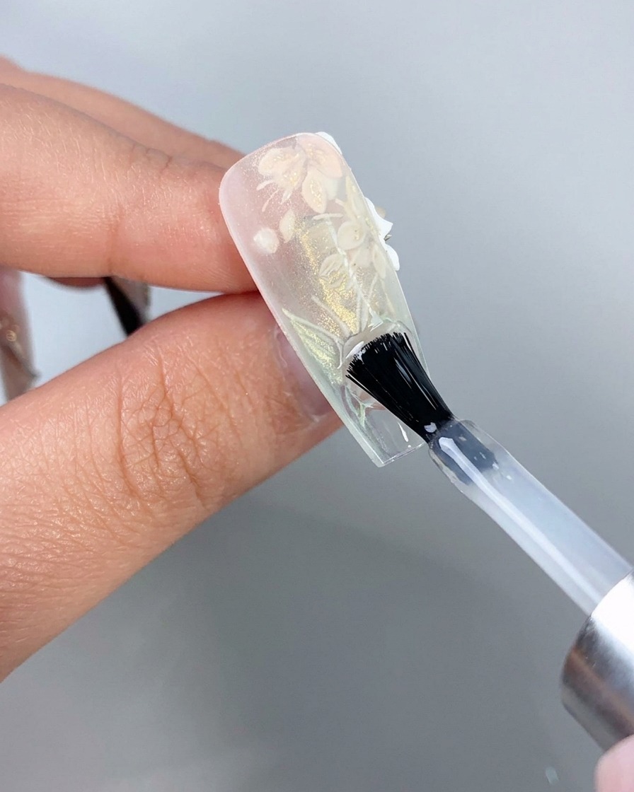 Un pincel de uñas aplicando una capa superior transparente en la parte inferior del borde libre de una punta de uña de extensión transparente.
