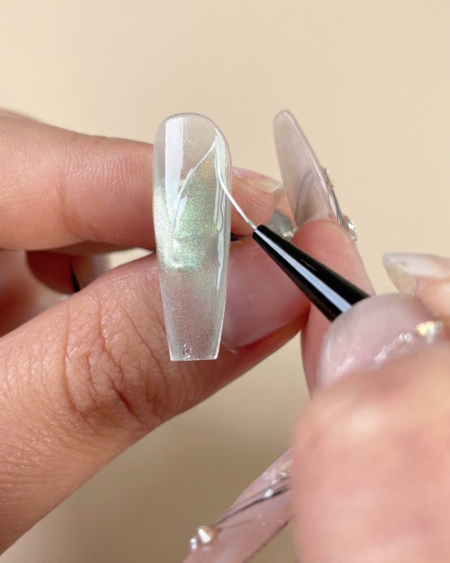 Un pincel delineador fino que dibuja líneas de venas blancas en una uña de gel de ojo de gato verde.