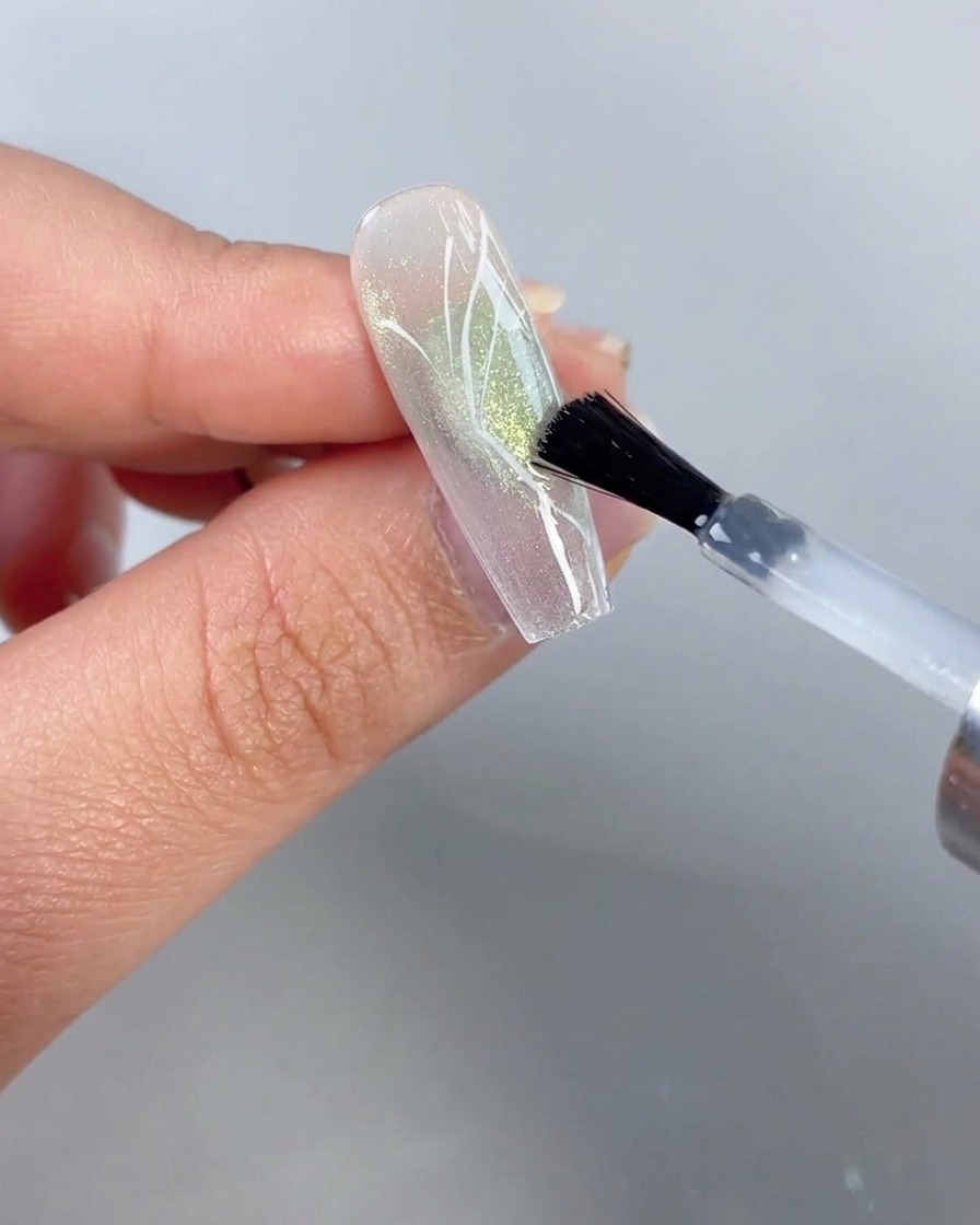 Un pincel aplicando una capa superior transparente sobre una uña con un diseño de media mariposa pintado contra un fondo de ojo de gato verde translúcido.
