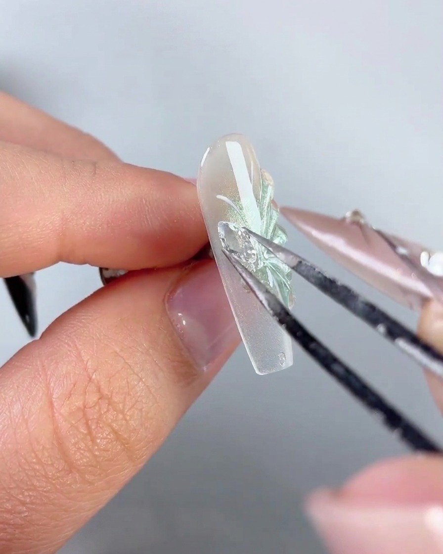 Un par de pinzas de metal colocando un dije brillante de ala de mariposa de pedrería en el lateral de una uña decorada.