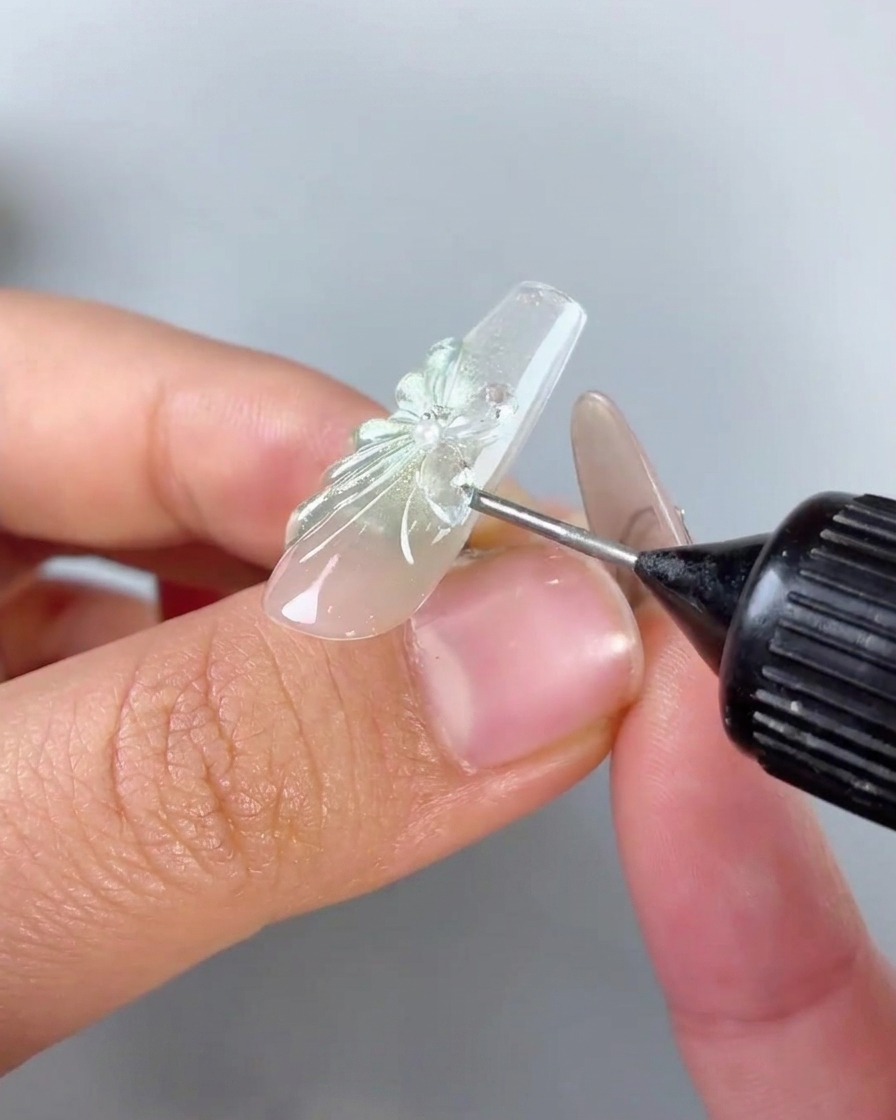 Una botella exprimible con punta de aguja aplicando gel transparente de relleno de espacios alrededor de la base y los bordes de un dije de mariposa de pedrería en una uña.