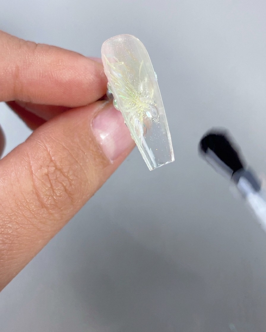 Un primer plano de una mano sosteniendo una uña transparente de estilo hielo con un diseño floral 3D boca abajo, mientras un pincel aplica capa superior transparente en la parte inferior del borde libre.