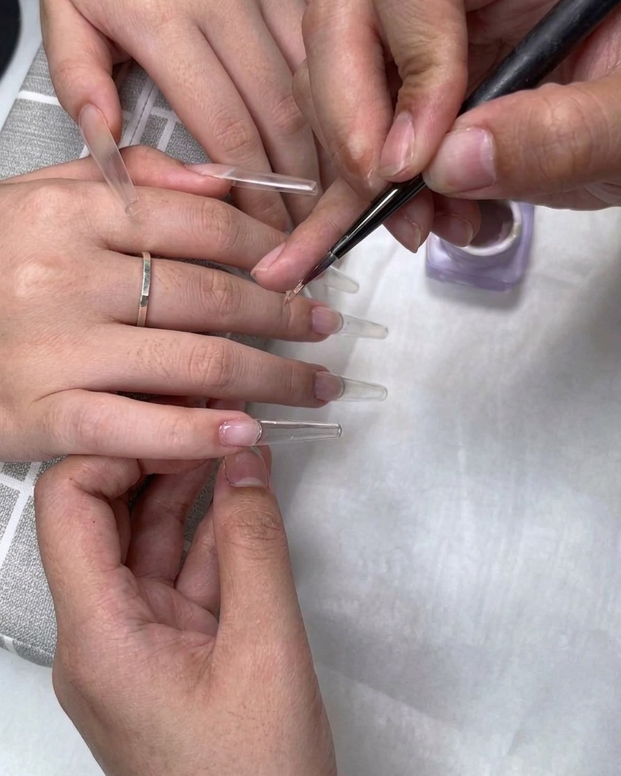 Un pincel fino aplicando esmalte de gel nude color té con leche cerca de la cutícula en una extensión de uña transparente.