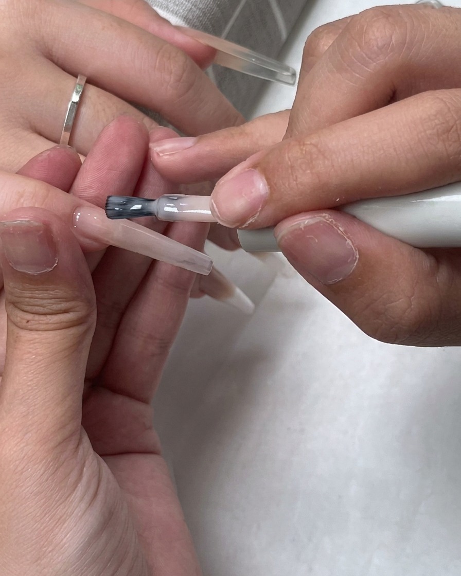 Un pincel aplicando una capa de esmalte nude translúcido sobre toda la superficie de la uña.