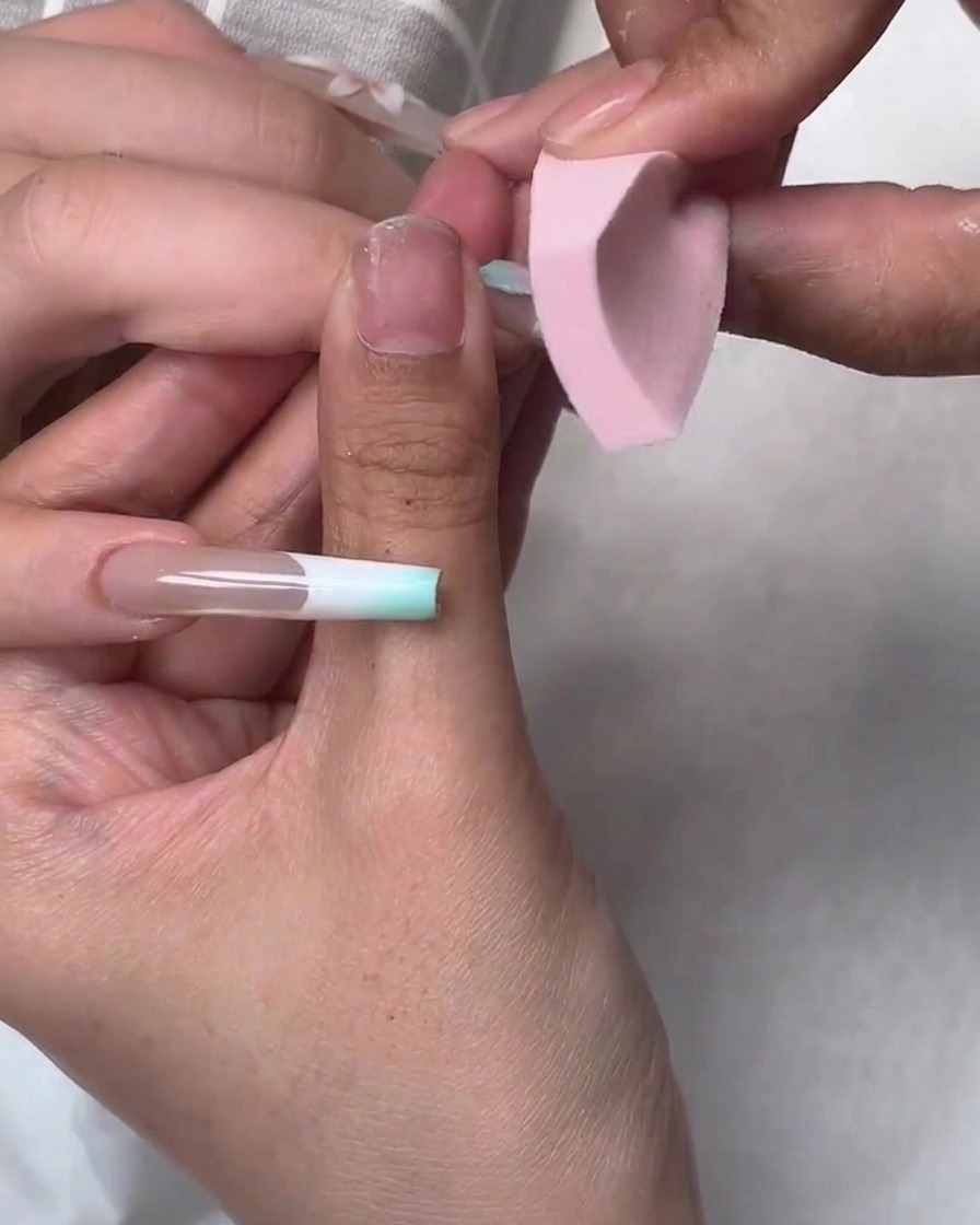 Una pequeña esponja rosa aplicando a toquecitos esmalte de gel azul en los bordes de una uña durante un tutorial de nail art.