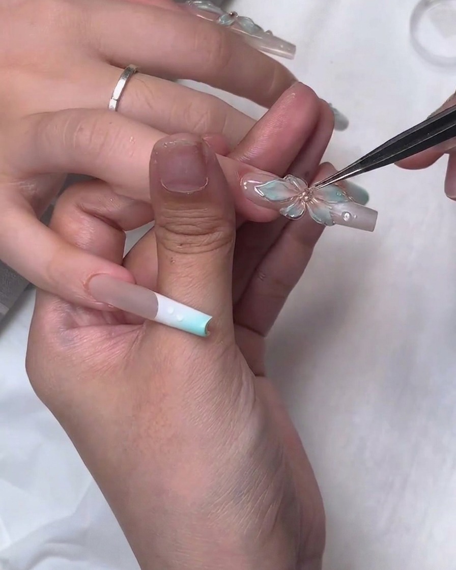 Un primer plano que muestra unas pinzas finas colocando con precisión un pequeño diamante de imitación en el centro de una flor de gel translúcido esculpida en 3D en una uña larga y cuadrada.